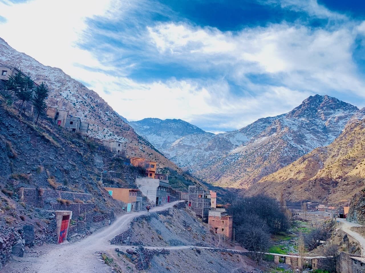A dirt track winds past a Berber village towards snow-capped peaks in the High Atlas